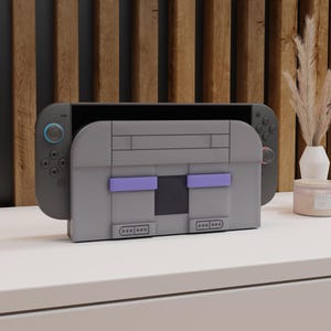 以下が含まれることがあります： レトロなゲーム機に似せてデザインされた、紫色のアクセントが付いたグレーのコンソールケースが、黒い携帯型ゲーム機と一緒に表示されています。 ケースは長方形で、ボタンの詳細があり、白い表面に配置されています。