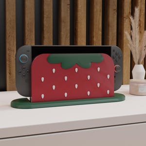 Könnte beinhalten: Eine dunkelgraue Spielkonsole in einem erdbeerförmigen roten Ständer mit grünem Sockel und Blattdetails. Die Erdbeere ist mit weißen, samenartigen Markierungen versehen. Die Konsole hat schwarze Controller mit blauen und rosafarbenen Akzenten.