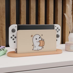 May include: A peach-colored Nintendo Switch dock with a cartoon ghost holding a pumpkin. The dock holds the Switch console and two white controllers. The design is on a white surface with a wooden background.