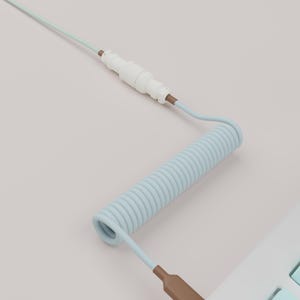 May include: A light blue coiled keyboard cable with a white connector and a brown USB-A connector. The cable is connected to a white keyboard with light blue keys. The background is a solid light pink.