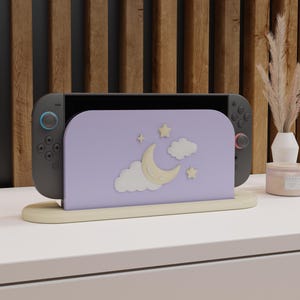 May include: A grey games console with blue and red controllers sits in a light purple stand. The stand has a crescent moon, stars, and clouds design. The stand is on a beige base, against a wooden background.