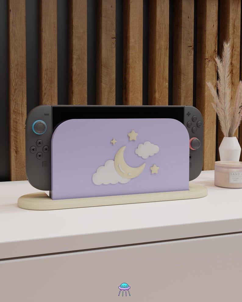 May include: A gray game console with blue and red controllers is placed in a light purple stand. The stand features a crescent moon, stars, and clouds design. The stand is on a beige base, set against a wooden background.