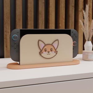 May include: A gray Nintendo Switch console rests in a peach-colored stand with a fox face design. The stand is on a white surface, with a wooden slatted background. A small vase and candle are visible.