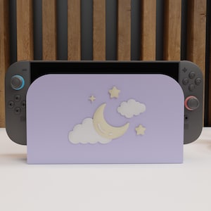 May include: A gray Nintendo Switch console with a lavender stand featuring a crescent moon, stars, and clouds. The stand is designed to hold the console upright. The background has a wooden slat design.