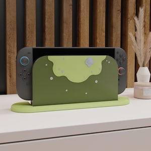 May include: A dark gray gaming console docked in a custom olive green stand. The stand features a light green scalloped top layer, a small light blue flower accent, and scattered white dots. The console has black controllers with blue and red accents.