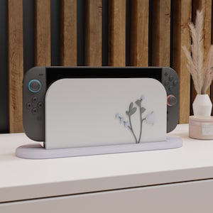 May include: A grey Nintendo Switch console in a white stand with a floral design. The stand sits on a white surface, with a wooden slatted background. A vase with dried flowers and a candle are in the background.