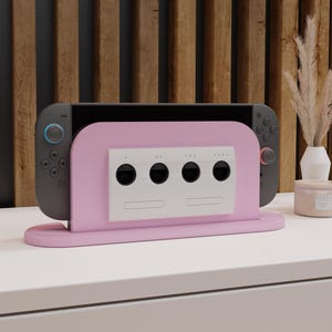 Peut inclure: Un support de console de jeu rose et blanc au design rétro. Le support contient une console de jeu grise avec des boutons bleus et rouges. Le support a un panneau blanc avec quatre cercles noirs et deux fentes rectangulaires. Le support est sur une surface blanche.