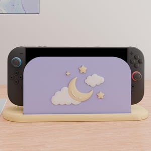 May include: A black gaming console docked in a light purple stand. The stand features a raised, cream-colored crescent moon, two white clouds, and two small stars. The console has a blue joystick on the left and a red joystick on the right.
