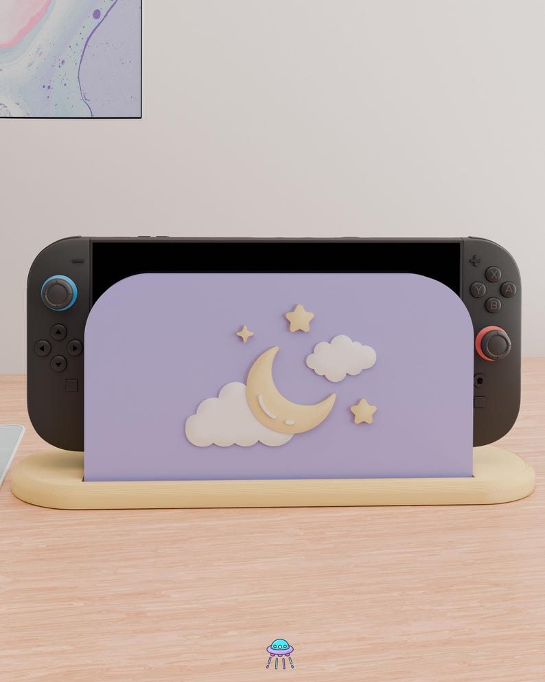 May include: A black gaming console docked in a light purple stand. The stand features a raised, cream-colored crescent moon, two white clouds, and two small stars. The console has a blue joystick on the left and a red joystick on the right.