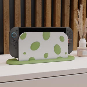 Może przedstawiać: Szara konsola Nintendo Switch jest prezentowana w zielonej podstawce z białą osłoną z zielonymi owalnymi wzorami. Podstawka i osłona zapewniają dekoracyjne i funkcjonalne wyświetlanie urządzenia do gier. Tło obejmuje drewniany panel i białą powierzchnię.
