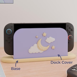 May include: A lavender gaming console dock cover featuring a raised cream-colored crescent moon, two clouds, and two stars. The cover sits in a light yellow base. The text "Base" and "Dock Cover" with arrows point to the respective parts.