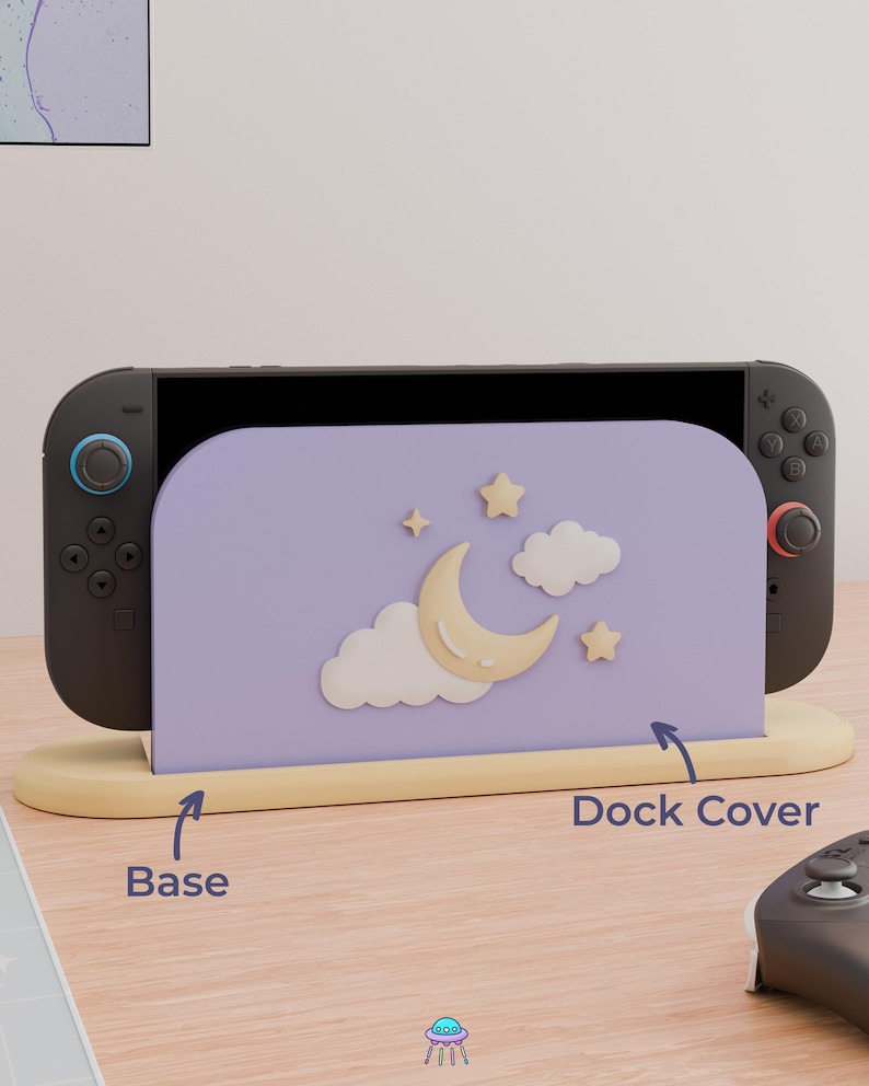 May include: A lavender gaming console dock cover featuring a raised cream-colored crescent moon, two clouds, and two stars. The cover sits in a light yellow base. The text "Base" and "Dock Cover" with arrows point to the respective parts.