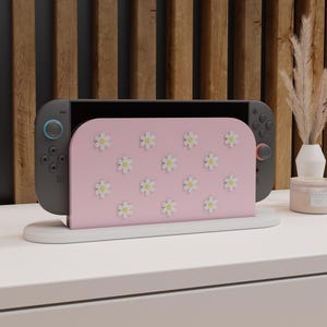Könnte beinhalten: Eine Nintendo Switch-Konsole mit einem rosa Dock mit weißen Gänseblümchen-Akzenten. Die Konsole ist grau mit blauen und roten Controllern. Das Dock steht auf einem weißen Ständer vor einem Holzpaneel-Hintergrund.