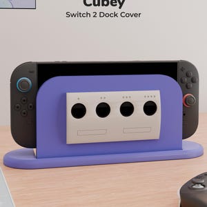 Può includere: Una cover per dock viola Cubey Switch 2 con un pannello frontale grigio chiaro dotato di quattro porte circolari per controller. La cover è posizionata sopra una console di gioco nera con accenti blu e rossi sui joystick, riportando il testo Cubey Switch 2 Dock Cover.