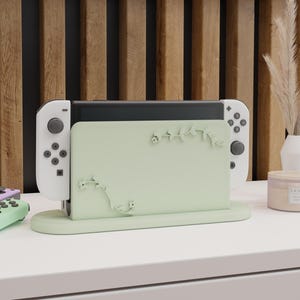 May include: A green and white Nintendo Switch console stand with a floral design. The stand has a small base and a raised platform for the console. The console is white with black buttons and a green and white Joy-Con controller.