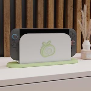 May include: A gray Nintendo Switch console docked in a light green stand with a white face featuring a green apple design. The stand sits on a white surface, with a wooden panel background.