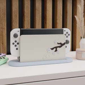 Puede incluir: Una base de carga para consola de videojuegos en color beige claro con un diseño de rama de árbol marrón oscuro y flores de cerezo blancas. La base se asienta sobre un soporte ovalado azul claro y sostiene una consola portátil blanca.