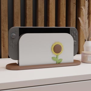 Puede incluir: Una consola de juegos gris descansa en un soporte marrón y blanco. El soporte presenta un diseño de girasol. La consola está colocada sobre una superficie blanca, con una pared de listones de madera en el fondo. También se ven una vela y un jarrón.