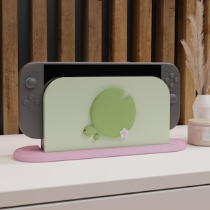 Puede incluir: Un soporte para Nintendo Switch verde claro y rosa con un diseño de nenúfar. El soporte presenta una parte trasera verde claro con un gran nenúfar verde y una pequeña flor rosa. La base es rosa y la Switch es gris.
