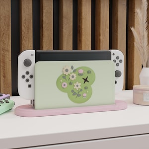 Könnte beinhalten: Eine Spielekonsolen-Dockingstation mit einer hellgrünen Frontplatte, die mit einem erhabenen Blumenmuster in Creme-, Rosa- und Dunkelgrüntönen verziert ist. Die Station steht auf einer blassrosa Basis und hält eine Konsole mit weißen Controllern.