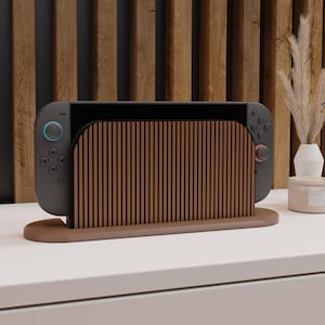 May include: A dark gray gaming console dock featuring a vertical slatted wood front panel and a smooth, oval-shaped wooden base. The console is inserted into the dock, displaying a black screen, with a blue joystick on the left and a red joystick on the right.
