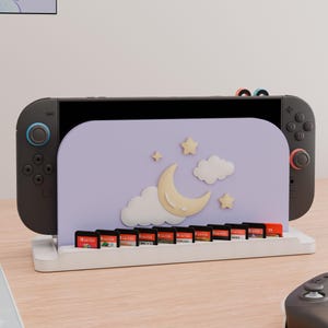 May include: A black gaming console docked in a white stand featuring a light purple panel decorated with a cream-colored crescent moon, white clouds, and small stars. The stand includes a row of slots holding ten small rectangular game cartridges.