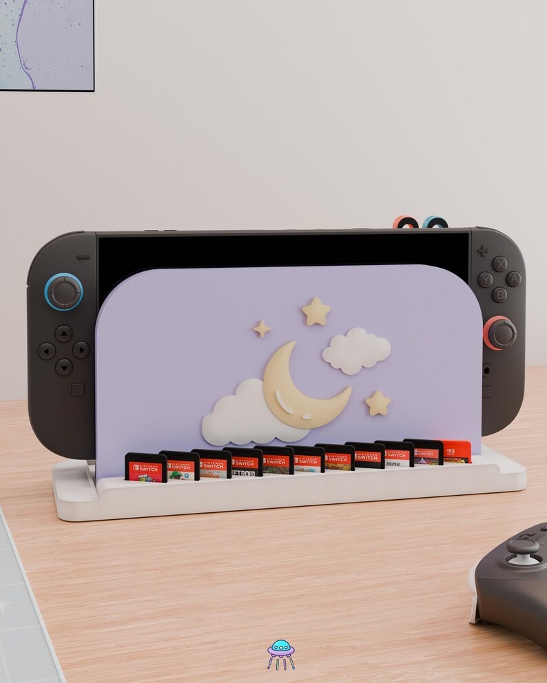 May include: A black gaming console docked in a white stand featuring a light purple panel decorated with a cream-colored crescent moon, white clouds, and small stars. The stand includes a row of slots holding ten small rectangular game cartridges.