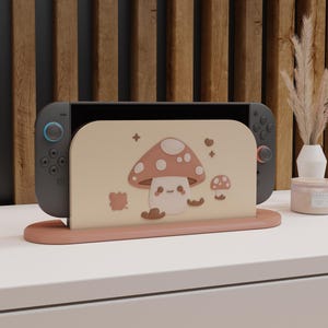 Könnte beinhalten: Eine graue Nintendo Switch-Konsole mit blauen und roten Controllern wird in einem rosa-beigen Ständer im Pilz-Design präsentiert. Der Ständer zeigt eine große Pilz-Illustration und eine kleine Pilz-Grafik.