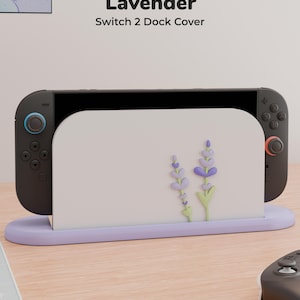 Può includere: Una cover per dock Switch 2 bianca con una base color lavanda e una grafica floreale lavanda. Il testo Lavender Switch 2 Dock Cover è visibile sopra il dispositivo. Il dock sostiene una console di gioco nera con joystick blu e arancioni.