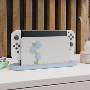 Könnte beinhalten: Weißer Nintendo Switch Konsolenständer mit blauer Basis und einem Blumendesign mit blauen und grünen Blumen. Der Ständer steht auf einer weißen Oberfläche.
