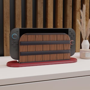 Puede incluir: Una consola Nintendo Switch con una base de estilo madera. La base es marrón con bandas negras y se asienta sobre una base roja. La consola es gris con botones azules y rojos.