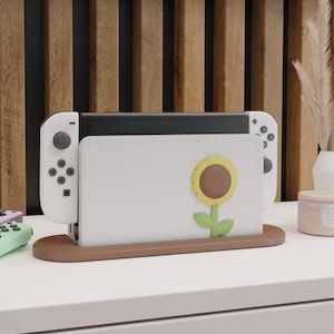 Puede incluir: Soporte de consola Nintendo Switch blanco con un diseño de girasol. El soporte tiene una base marrón y una parte superior blanca con un hueco para la consola. El soporte está diseñado para sostener la consola y dos mandos.