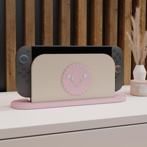 以下が含まれることがあります： 青と赤のコントローラーが付いたグレーのゲーム機が、ピンク色のスタンドにドッキングされています。スタンドには、ピンクの花柄のデザインが施されたベージュのインサートがあります。白い表面に木製の背景があります。