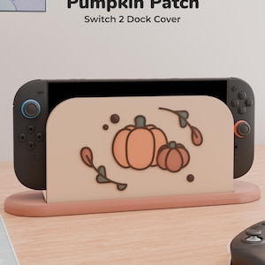 Könnte beinhalten: Eine beige Switch 2 Dockingstation-Abdeckung mit einer Grafik aus zwei orangefarbenen Kürbissen und braunen Blattzweigen. Der Text Pumpkin Patch Switch 2 Dock Cover steht darüber. Die Station ist mit einer schwarzen Spielkonsole verbunden.
