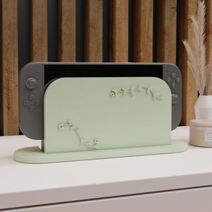 Könnte beinhalten: Ein hellgrüner Nintendo Switch-Dock mit floralem Design. Der Dock ist so konzipiert, dass er die Switch-Konsole aufrecht hält. Der Hintergrund umfasst eine Holzverkleidung und eine weiße Oberfläche.