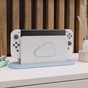 May include: A white Nintendo Switch console with attached controllers is docked in a light blue stand. The stand has a cloud design. The console is on a white surface with a wooden slatted background.