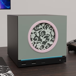 May include: A square computer case with a matte black frame and a sage green front panel. The centre features a circular vent with a light pink rim and an intricate white floral filigree pattern. A thin blue light strip sits above the black base.