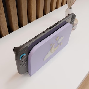 May include: A Nintendo Switch console with a lavender-colored stand featuring a whimsical design of a crescent moon, clouds, and stars. The console is gray and black, with blue accents on the controller.
