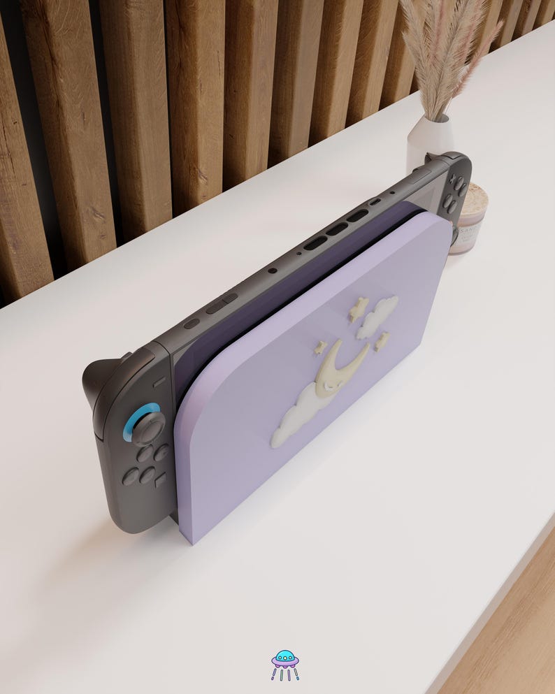 May include: A Nintendo Switch console with a lavender-colored stand featuring a whimsical design of a crescent moon, clouds, and stars. The console is gray and black, with blue accents on the controller.
