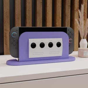 May include: A gray and black handheld gaming console is displayed in a purple stand. The stand has a silver GameCube-style faceplate with four circular holes. The stand is on a white surface with a wooden background.