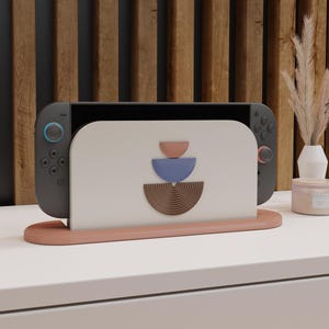 May include: A gray Nintendo Switch console is placed in a peach-colored stand. The stand has a cream-colored back with a geometric design in peach, blue, and brown. The stand is on a white surface.