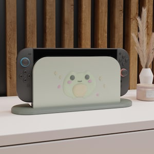 May include: A gray Nintendo Switch console with blue and red controllers is docked in a light green stand. The stand features a cute frog design. The setup is on a white surface with a wooden background.