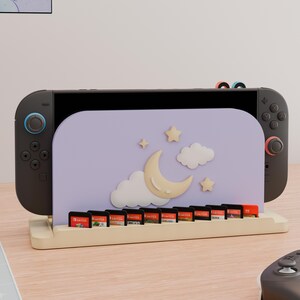 May include: A light purple gaming console stand featuring a crescent moon, clouds, and stars design. The stand holds a black gaming console and includes a row of ten small rectangular game cartridges in a light-colored wooden base.