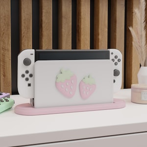 May include: A white Nintendo Switch console with attached controllers, set in a pink stand. The console's screen features two pink strawberry designs. The stand and console are on a white surface, with a wooden slat background.