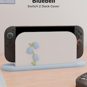 Peut inclure: Un cache pour station d'accueil Switch 2 blanc orné d'un motif de fleurs de campanules bleu clair avec des tiges vertes. Le cache repose sur une base bleu clair et contient une console de jeu noire avec des joysticks bleu et rouge. Texte Bluebell.