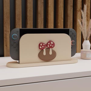 Puede incluir: Soporte de madera beige claro para Nintendo Switch con diseño de setas. El soporte sostiene una consola Nintendo Switch gris. Las setas son rojas con lunares blancos y se asientan sobre una base marrón. El soporte está sobre una superficie blanca.