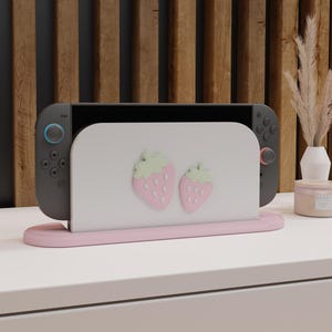 以下が含まれることがあります： ピンクと白のNintendo Switchスタンド。イチゴのデザインが2つあります。スタンドはSwitchコンソールを保持するように設計されています。イチゴはピンク色で、緑色の葉が付いています。スタンドはピンク色のベースと白い背面パネルを備えています。