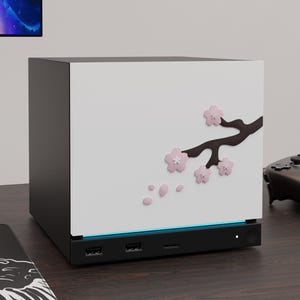 May include: A black and white computer case featuring a decorative dark brown tree branch with light pink cherry blossom flowers and falling petals on the side panel. The base includes two USB ports, a central port, and a small circular power button.