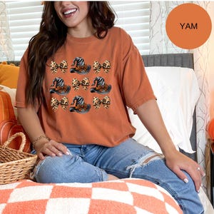 May include: A burnt orange short-sleeve t-shirt featuring a repeating graphic pattern of black witch hats with orange bands and leopard print bows. The shirt has a crew neckline and a relaxed fit, showcasing a seasonal design with festive motifs.