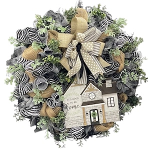 May include: A decorative wreath featuring black and white striped mesh, tan burlap loops, and green foliage. A central bow combines tan burlap, polka dot, and striped ribbons. A wooden house sign reads IT'S GOOD TO BE home with a small wreath on the door.